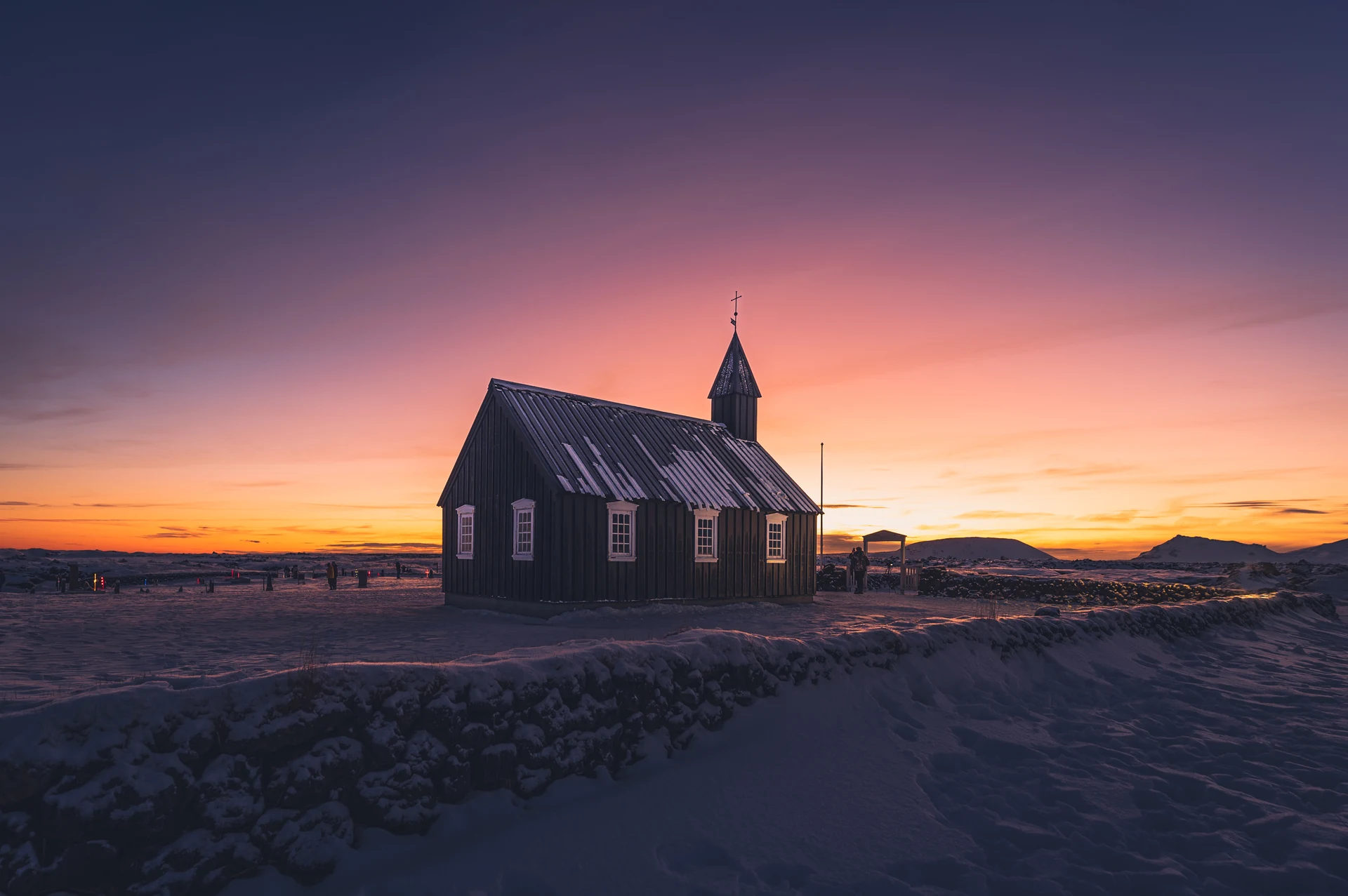Une petite église sombre, dans la neige, devant un ciel orange et violet.