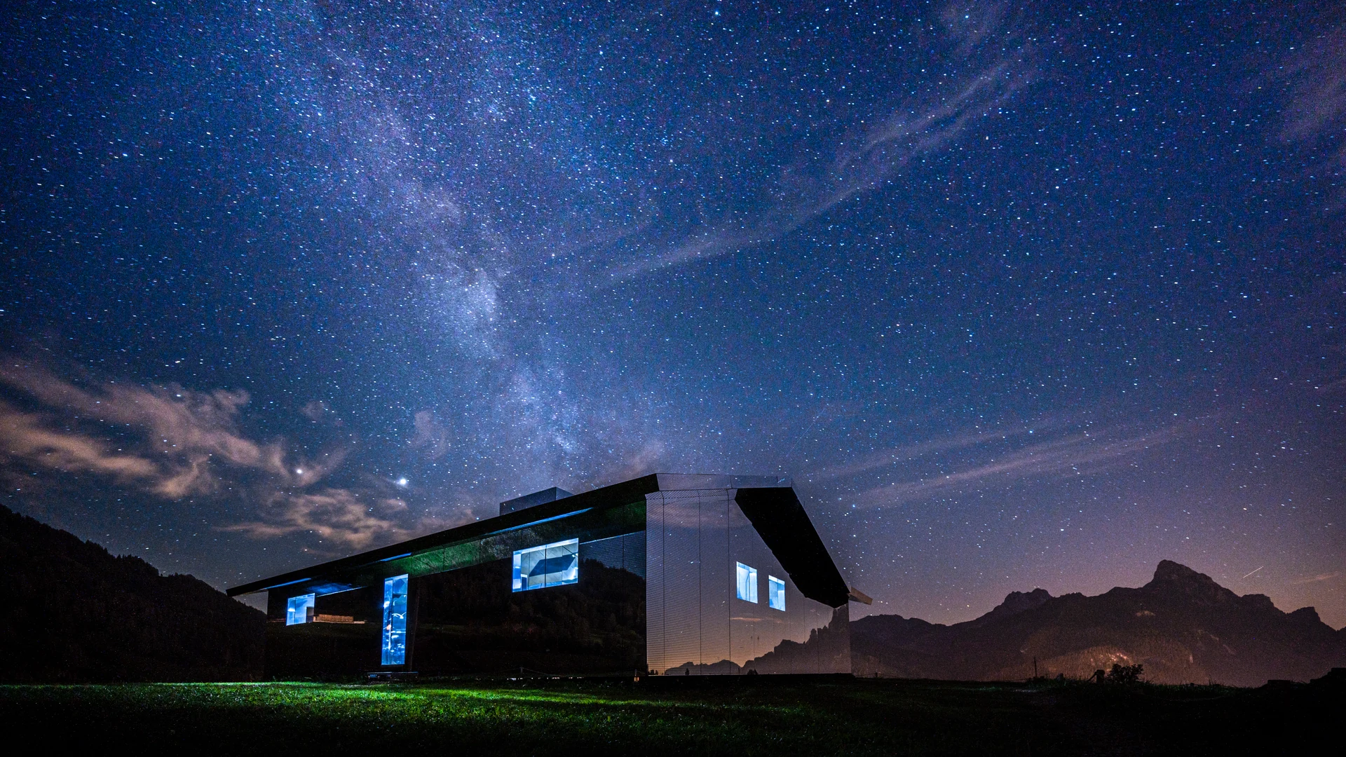 Une maison éclairée sous la Voie lactée et les silhouettes des montagnes.